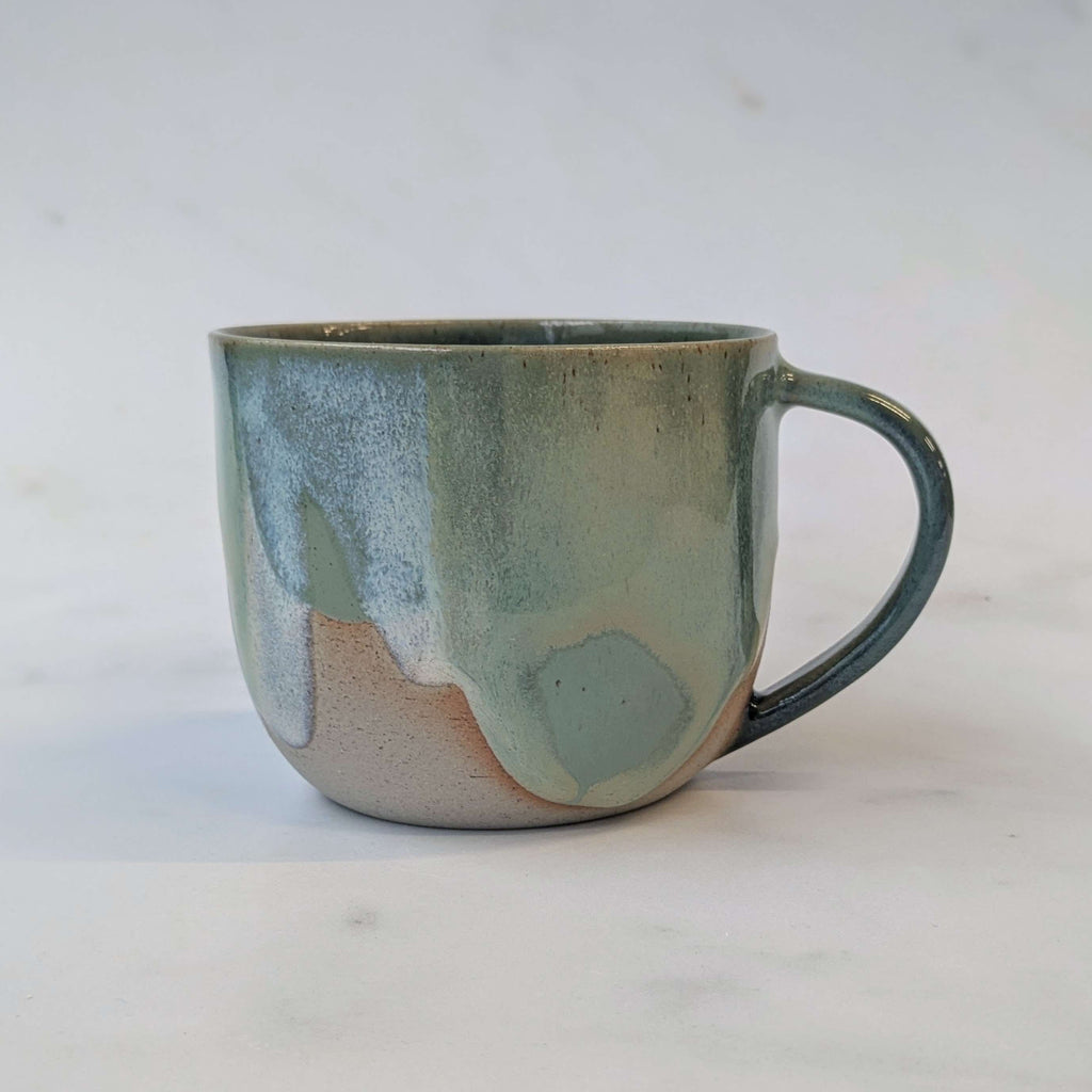 A handmade ceramic mug with a rounded shape, a C-shaped handle, and a textured glaze featuring a gradient of sage green, slate blue, and a warm beige base, set against a light off-white background.