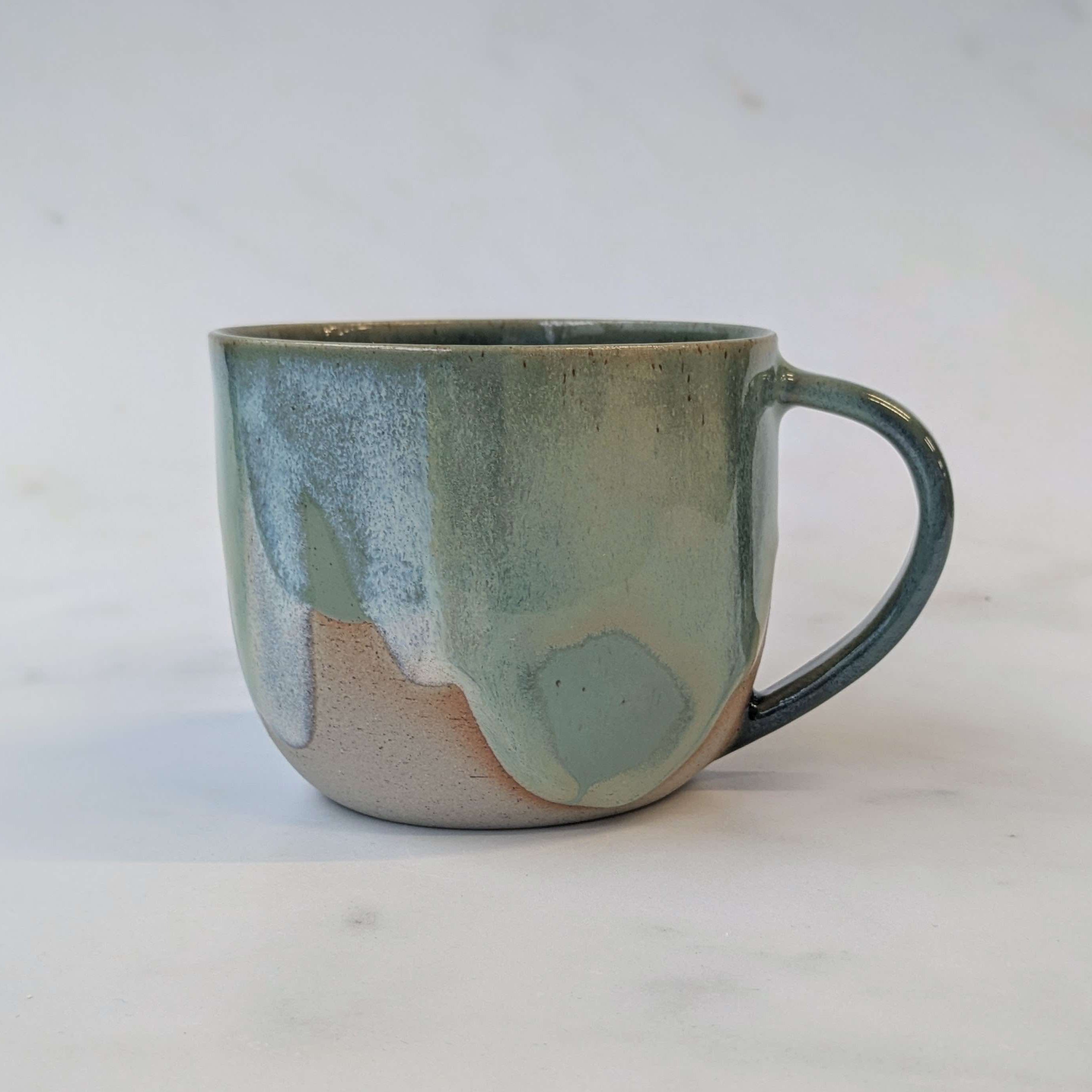 A handmade ceramic mug with a rounded shape, a C-shaped handle, and a textured glaze featuring a gradient of sage green, slate blue, and a warm beige base, set against a light off-white background.