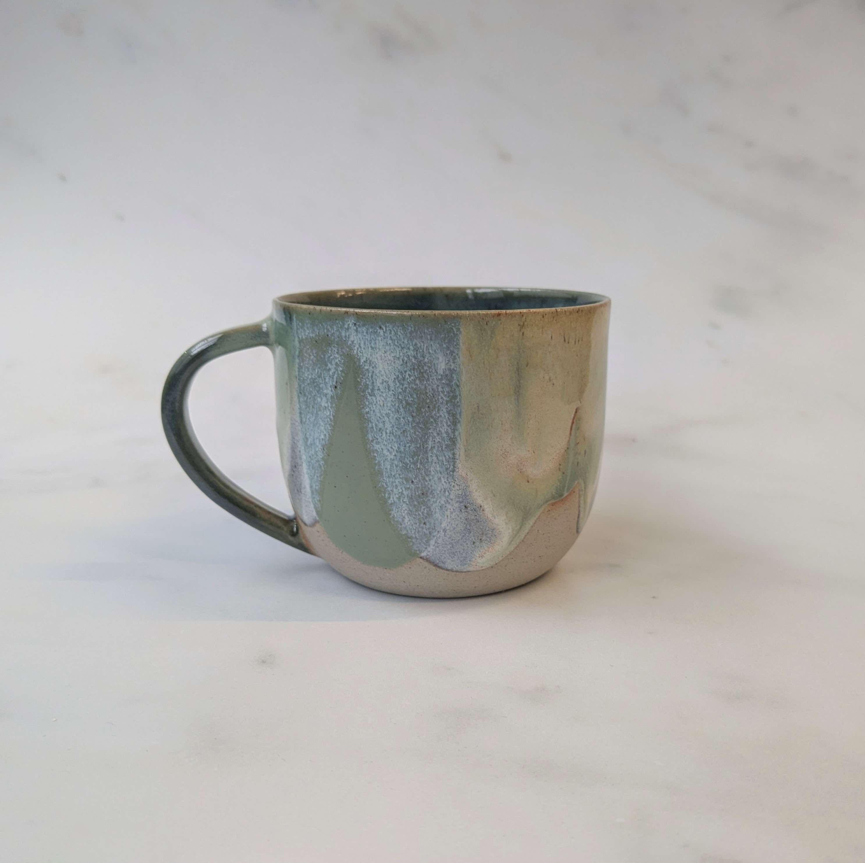 A handmade ceramic mug with a marbled, fluid glaze in shades of sage green, dark blue, and warm beige, set against a light marble background.