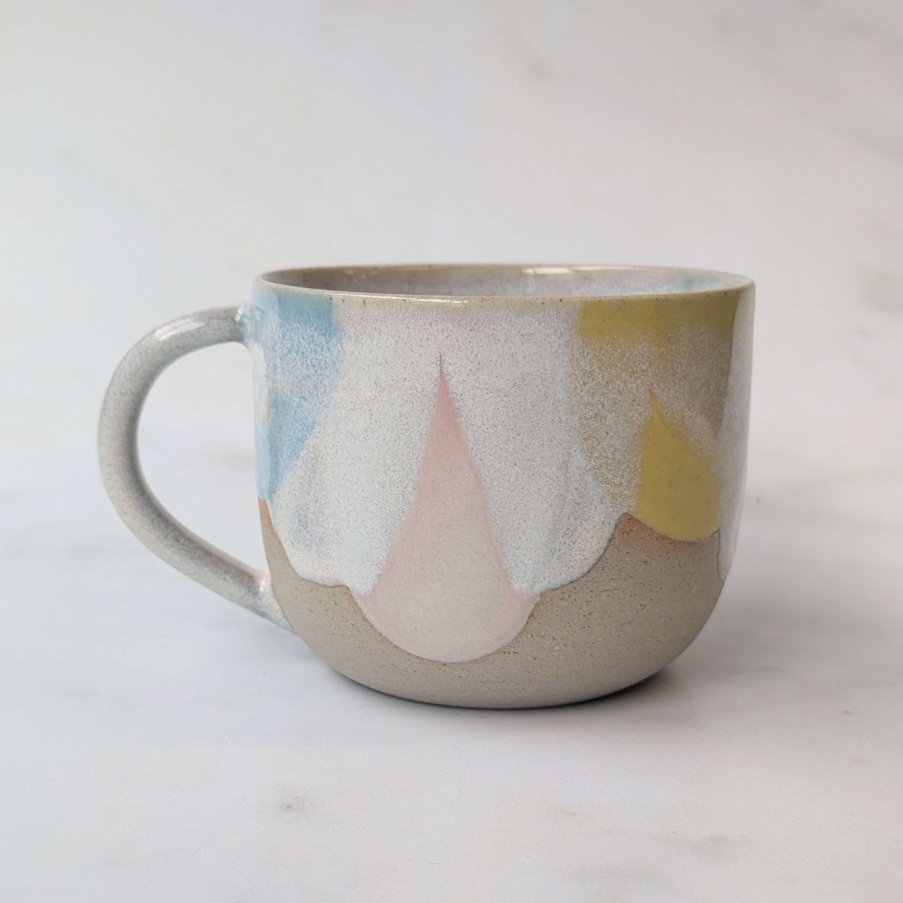 A handmade ceramic mug with a speckled gray and beige base, featuring a dripping glaze design in pastel shades of blue, pink, and yellow, set against a light marble background.